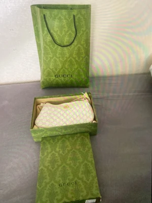 Gucci beige axelremsväska monogram - Säljer en snygg beige axelremsväska från Gucci med klassiskt GG-monogrammönster och guldfärgad logga framtill. Väskan har en mjuk, rundad form och är tillverkad i canvas med läderdetaljer. Kommer med original dustbag, box och påse.