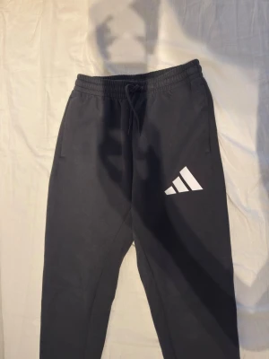 Svarta mjukisbyxor från Adidas - Säljer ett par svarta mjukisbyxor från Adidas med vit logga på vänster ben. Byxorna har dragsko i midjan och sidofickor. Tillverkade i mjukt och bekvämt material, perfekta för chill eller träning.