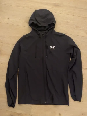 Svart vindjacka från Under Armour - En snygg under armour jacka i bra skick. Jackan är inte bara i bra skick utan passar även till nästan allt och alla. Är en jacka perfekt för att skydda mot vindar samtidigt hålla dig både varm och snygg.