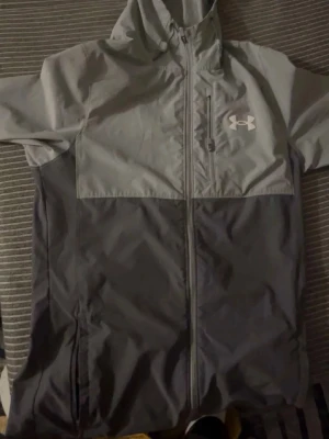 Grå vindjacka från Under Armour - Snygg tvåfärgad vindjacka från Under Armour i ljusgrått och mörkgrått. Jackan har huva, hel dragkedja framtill och sidofickor med dragkedja. Tillverkad i lätt och vindtätt material, perfekt för blåsiga dagar. Under Armour-logga på bröstet.