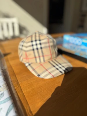 Beige Burberry keps med rutigt mönster - Säljer en klassisk beige keps från Burberry med det ikoniska rutiga mönstret i svart, vitt och rött. Kepsen har böjd skärm och justerbar rem bak. Perfekt accessoar för dig som vill ha en tidlös och trendig look.