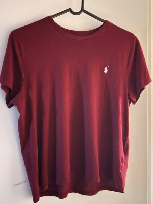 Vinröd t-shirt från Polo Ralph Lauren - Snygg vinröd t-shirt från Polo Ralph Lauren med klassisk rund hals och korta ärmar. Tillverkad i mjuk bomull och prydd med den ikoniska broderade loggan på bröstet. Perfekt basic-plagg för en clean och stilren look.