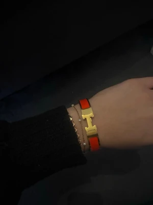 Orange armband med H-spänne - Snyggt armband i orange och ”guld” med det ikoniska H-spännet. Armbandet är i metall med emaljdetaljer och har en stilren, modern design som sticker ut. Perfekt accessoar för att lyfta vilken outfit som helst.
