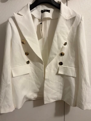 Vit kavaj med guldknappar  - Stilren vit kavaj från FLY med snygga guldknappar framtill och klassisk krage. Kavajen har två fickor med lock och en rak passform som ger en clean look. Perfekt för dig som vill sticka ut med en fräsch och modern stil./// Den är dock lite mindre så storleken skulle nog anses vara L 