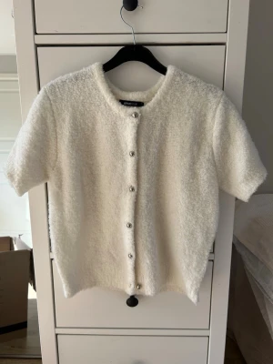 Vit kortärmad kofta från Gina Tricot - Mysig vit kofta från Gina Tricot med korta ärmar och rund halsringning. Koftan har en fluffig, mjuk textur och knäpps med silvriga knappar framtill. Perfekt för lager-på-lager och en clean look.