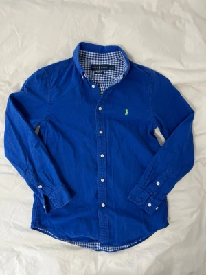 Ralph Lauren skjorta - Snygg blå skjorta från Ralph Lauren med klassisk button-down krage och vita knappar. Insidan av kragen har ett rutigt mönster i blått och vitt. Ikoniska gröna loggan broderad på bröstet. Tillverkad i mjuk bomull och har lång ärm. Storlek L(14-16) så sitter ungefär som S
