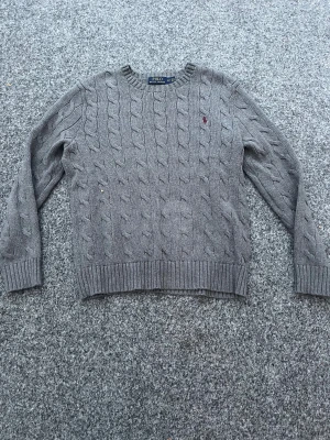Grå kabelstickad tröja Polo Ralph Lauren - Snygg grå kabelstickad tröja från Polo Ralph Lauren med rund hals och ribbade muddar. Tröjan har ett broderat rött Polo-logo på bröstet och är långärmad. Perfekt för vintern.