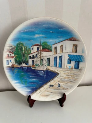 Dekorativ tallrik med hamnmotiv - Dekorativ rund tallrik med handmålat motiv av en hamn, vita hus med blå detaljer, båtar och träd. Tallriken har blå, vita och gröna färger och står på ett mörkbrunt ställ. Perfekt som prydnad eller väggdekoration.Diameter 24 cm 