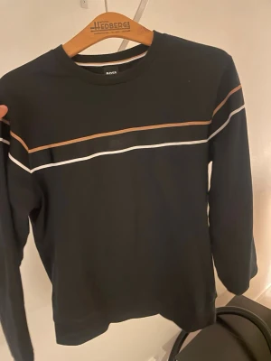 Svart tröja med ränder från BOSS - Svart långärmad tröja från BOSS med tunna horisontella ränder i vitt och beige över bröstet och ärmarna. Rund halsringning och diskret BOSS-logga framtill. Perfekt för en stilren och avslappnad look.
