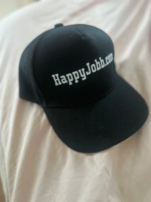 Svart keps med HappyJobb.com-tryck - Cool svart keps med böjd skärm och vit text framtill där det står HappyJobb.com. Klassisk modell med justerbar baksida och ventilerande hål. Perfekt accessoar för en avslappnad stil.