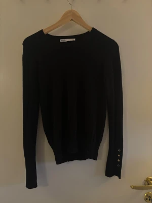 Svart stickad tröja från Zara - En svart, tunnstickad tröja från Zara med rund halsringning och långa ärmar. Tröjan har dekorativa knappar vid ärmsluten som ger en snygg detalj. Perfekt att matcha med jeans eller kjol för en clean look.Hör av dig om du har några frågor! 💗🫶🏼