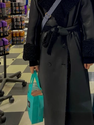 Svart lång fuskpäls kappa - Snygg svart lång kappa med fuskpälsdetaljer på krage och ärmslut. Kappan har dubbelknäppning, bälte i midjan och två fickor framtill. Materialet är glansigt och ger en lyxig känsla. Perfekt för kalla dagar när du vill sticka ut.