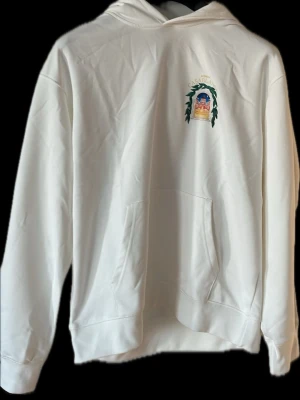 Vit hoodie med Casablanca-tryck - Säljer en vit hoodie från Avenida med ett färgglatt tryck på bröstet där det står 'CASABLANCA' och motiv av en byggnad omgiven av gröna blad. Hoodien har huva och stor magficka framtill. Perfekt för dig som gillar stilrena plagg med unik detalj.