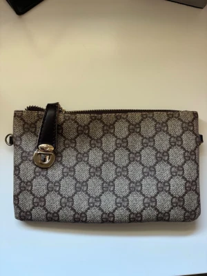Brun monogram kuvertväska - Snygg kuvertväska i beige och brunt med klassiskt GG-monogram. Väskan har dragkedja upptill, detaljer i mörkbrunt läder och en silverfärgad spänndetalj. Tillverkad i canvas med läderdetaljer och har en extra ficka med dragkedja på baksidan.