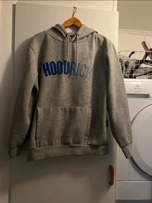 Grå HOODRICH hoodie med blå text - Snygg grå hoodie från HOODRICH med stor blå broderad text på bröstet. Klassisk känguruficka framtill, justerbar huva med dragsko och ribbade muddar. Perfekt för en avslappnad streetwear-look. Mjuk och skön bomullsblandning.