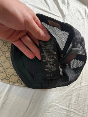 Gucci beige GG Supreme keps - Snygg beige keps från Gucci med klassiskt GG Supreme-mönster och svart mesh på baksidan. Justerbar rem med grön-röd randig detalj. Tillverkad i Italien och har en modern, avslappnad passform. Perfekt för dig som vill ha en ikonisk och trendig accessoar.
