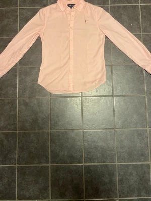 Ljusrosa skjorta från Ralph Lauren - Klassisk ljusrosa skjorta från Ralph Lauren med broderad logga på bröstet. Skjortan har lång ärm, knappar framtill och en tidlös, normal passform. Perfekt till både jeans och chinos för en clean look.