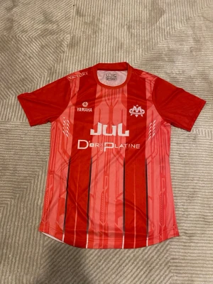Röd Marseille JUL fotbollströja - Snygg röd fotbollströja från C'est Marseille Bébé med vertikala svarta och ljusröda ränder. Tröjan har tryckta loggor från Yamaha och Jul samt texten D'or Platine. Tillverkad i lätt och ventilerande syntetmaterial, perfekt för träning eller match.