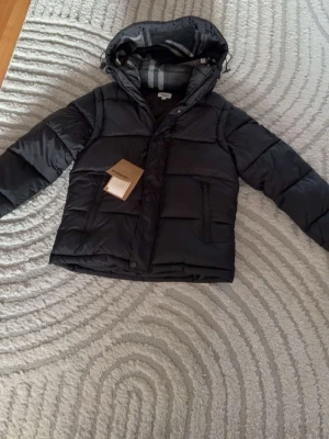 Svart dunväst med avtagbar huva - Snygg svart dunväst med avtagbar huva och dragkedja framtill. Västen har två stora fickor med dragkedja och rutigt innerfoder i huvan. Perfekt för lager-på-lager och funkar till många olika stilar.