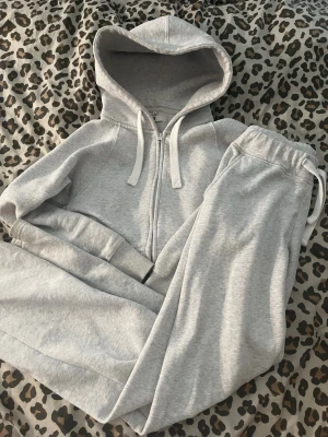 Grått mjukisset med hoodie - Säljer ett grått mjukisset med zip-hoodie och matchande byxor. Hoodien har dragkedja, fickor och snören i huvan. Byxorna har resår i midjan och snörning. Perfekt för chill dagar hemma eller när du vill vara bekväm.