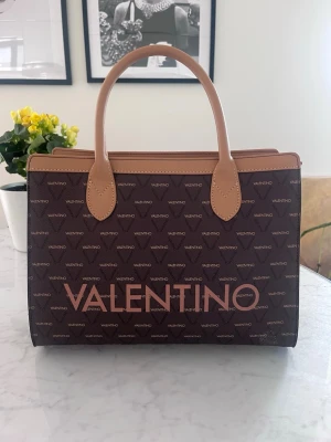 Brun handväska från Valentino - Snygg brun handväska från Valentino med ljusbruna handtag och detaljer. Väskan har ett mönster med Valentino-loggan över hela ytan och stor logotyp framtill. Stilren och rymlig modell i rektangulär form, perfekt för att bära dina essentials. Kommer med axelrem.