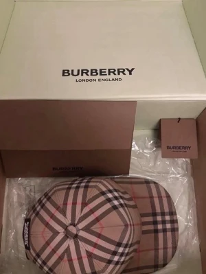 Burberry beige  keps - Säljer en snygg keps från Burberry med klassiskt beige rutmönster och detaljer i svart, vitt och rött. Kepsen har justerbar rem baktill och är tillverkad i ett slitstarkt tyg. Perfekt för dig som vill ha en ikonisk accessoar med London-vibe.