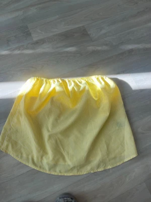 Gul topp med tunna ränder - Säljer en söt gul topp med tunna vita ränder. Kjolen har en enkel och luftig design med resår i midjan, perfekt för varma dagar. Materialet känns lätt och bekvämt, och den passar dig som gillar en fräsch och ungdomlig stil.