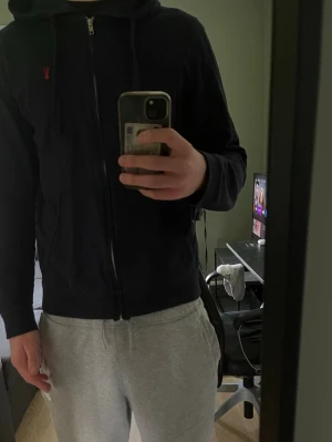 Zip Hoodie - Blå hoodie från Next med dragkedja framtill och två fickor. Klassisk huva med snören och en liten röd broderad logga på bröstet. Tillverkad i mjukt bomullsmaterial som är skönt att ha på sig. Perfekt för en avslappnad och stilren look.