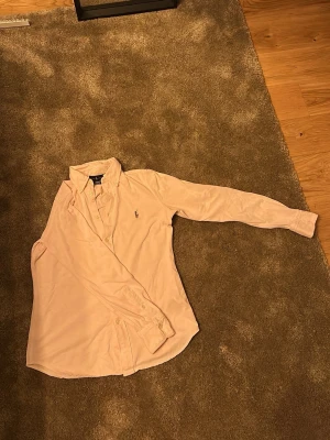 Rosa skjorta från Ralph Lauren - Klassisk rosa skjorta från Ralph Lauren med broderad logga på bröstet. Skjortan har lång ärm, knappar framtill och en tidlös krage. Perfekt för dig som gillar stilrena plagg med exklusiv känsla.