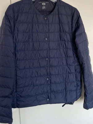 Mörkblå lättviktsdun jacka Uniqlo - Snygg mörkblå lättviktsdunv jacka från Uniqlo med rund halsringning och tryckknappar framtill. Västen har ett stilrent, quiltat mönster och är tillverkad i tunt, smidigt material som håller värmen utan att kännas klumpig. Perfekt för lager på lager under kyliga dagar.