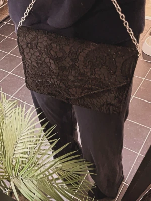 Svart spets väska - Snygg svart kuvertväska med spetsdetaljer och silverfärgad kedjerem. Väskan har magnetlås och insida i satinliknande material. Perfekt för dig som vill ha en elegant accessoar med lite extra attityd.