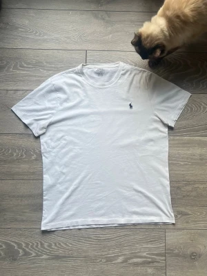 Vit Polo Ralph Lauren T-shirt – Storlek L – Custom Slim Fit - Klassisk vit t-shirt från Polo Ralph Lauren med den ikoniska mörkblå logotypen broderad på bröstet. En tidlös basfavorit som passar till allt. • Märke: Polo Ralph Lauren • Modell: Custom Slim Fit • Storlek: L (Large) • Färg: Vit • Skick: som ny 