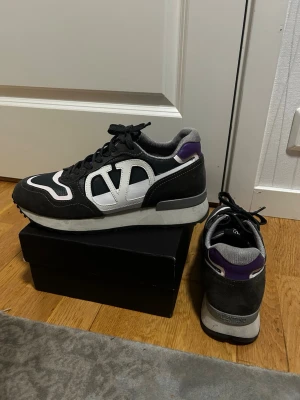 Valentino Garavani sneakers  - Snygga sneakers från Valentino Garavani i svart mocka och textil med vita och lila detaljer. Skorna har en tydlig V-logga på sidan, svart snörning och en gråvit sula. Perfekta för dig som gillar exklusiva och trendiga sneakers.