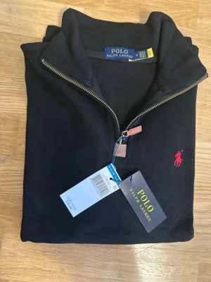 Svart halvzip från Polo Ralph Lauren - Svart långärmad tröja från Polo Ralph Lauren med halvzip och ståkrage. Klassisk röd broderad logga på bröstet och metallblixtlås med läderdetalj. Perfekt för en stilren och avslappnad look.