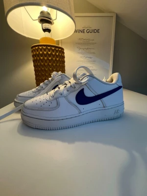 Nike Air Force 1 vita med blå swoosh - Klassiska Nike Air Force 1 sneakers i vitt läder med blå swoosh på sidan. Skorna har perforerad tå, vita skosnören och en robust sula med Air-dämpning. Perfekt för dig som gillar tidlös och sportig stil.