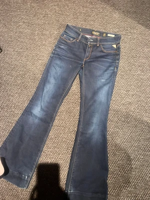 Blå bootcut jeans från Replay - Snygga blå bootcut jeans från Replay med klassiska fem fickor och kontrastsömmar. Jeansen har en lätt tvättad look och utsvängda ben. Tillverkade i denim med detaljer i metall och läderpatch på fickan.
