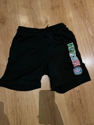 Svarta Nirvana shorts med tryck - Svarta shorts med färgglatt Nirvana-tryck på ena benet och rund logga. Resårmidja med snörning och bakficka. Perfekta för chill dagar eller konserter. Mjuk bomullskänsla och relaxed fit.