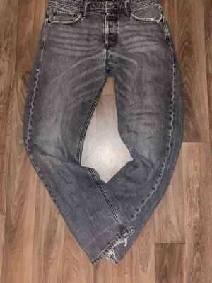 Grå tvättade raka jeans - Säljer ett par grå tvättade jeans med rak passform och klassisk femficksdesign. Jeansen har slitna detaljer vid benslut och en avslappnad look. Materialet är denim i bomull och färgen är en snygg grå nyans med tvättade effekter.