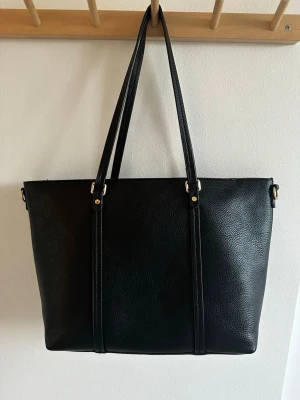 Svart handväska  -  svart  handväska med två långa axelband och guldfärgade detaljer. Väskan har rymligt huvudfack med dragkedja och innerficka för småsaker. Perfekt för dig som vill ha plats för både laptop och vardagsprylar. Perfekt till skolan. Väskan har några få slitningar på baksidan men syns inte när man har den på axeln 