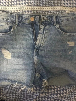 Blå jeansshorts  - Säljer ett par blå jeansshorts med rå kant och slitna detaljer framtill. Shortsen har klassisk femficksdesign, bälteshällor och dragkedja med knapp. Perfekta för en avslappnad och trendig look.