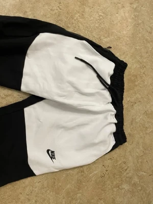 Nike svartvita mjukisbyxor - Säljer ett par svartvita mjukisbyxor från Nike med klassisk logga på benet. Byxorna har dragsko i midjan och elastiska muddar vid bensluten. Perfekta för chill dagar eller träning. Snygg colorblock-design som sticker ut.
