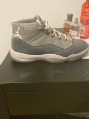 Gråa Air Jordan 11 sneakers - Säljer ett par gråa Air Jordan 11 sneakers med vita detaljer och vit sula. Skorna har högt skaft, klassisk Jumpman-logga på sidan och snörning framtill. Ovandelen är i en mix av läder och syntetmaterial med glansig finish på vissa partier.
