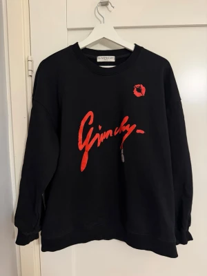 Svart Givenchy sweatshirt med röd logga - Svart sweatshirt från Givenchy med röd grafisk logga och tryck framtill samt Givenchy-symbol på ryggen. Oversized passform med rund halsringning och ribbade muddar. Tillverkad i mjuk bomullsblandning som känns skön mot huden. Perfekt för en trendig streetwear-look.