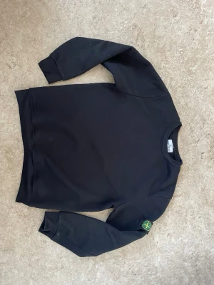 Svart sweatshirt från Stone Island - Säljer en svart sweatshirt från Stone Island med den klassiska logotypen på vänster ärm. Tröjan har rund halsringning och ribbade muddar vid ärmslut och nederkant. Perfekt för dig som gillar streetwear och vill ha en stilren look.