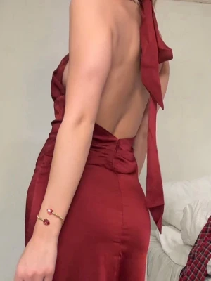 Balklänning - Säljer en vinröd långklänning i satin med halterneck och öppen rygg. Klänningen har en snygg slits framtill och knytdetalj i nacken. 