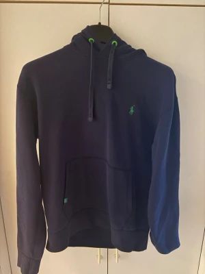 Mörkblå hoodie från Polo Ralph Lauren - Mörkblå hoodie från Polo Ralph Lauren med klassisk huva och dragsko med gröna detaljer. Broderad grön logga på bröstet och stor magficka framtill. Tillverkad i mjukt bomullsmaterial, perfekt för en avslappnad och stilren look.