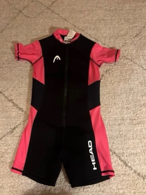 Svart och rosa våtdräkt HEAD - Säljer en svart och rosa våtdräkt från HEAD med korta ärmar och ben. Dräkten har dragkedja framtill och vita logotyper på bröstet och benet. Tillverkad i stretchigt funktionsmaterial som passar perfekt för simning och vattensport.
