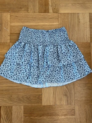 Blommig volangkjol i blått med blommor - Supersöt kort kjol med blommigt mönster i blått och vitt. Kjolen har bred resår i midjan och dubbla volanger. 