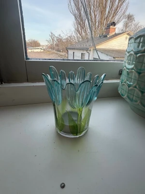 Blå glasvas med blomblad - Unik glasvas i genomskinligt glas med blå och gröna detaljer som liknar blomblad runt kanten. Perfekt som dekoration eller för mindre blommor. Vasen har en rund form och konstnärlig design som sticker ut i fönstret.