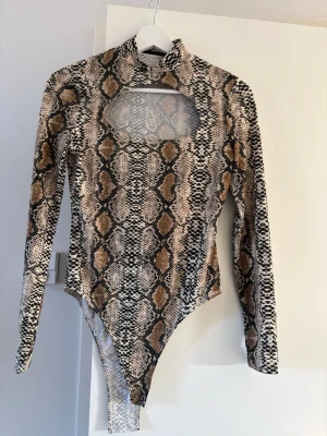 Ormmönstrad långärmad body - Snygg body med ormmönster i beige, brunt och svart. Långärmad modell med hög hals och en stor cut-out vid bröstet. Praktisk knäppning i grenen. Stretchigt material som sitter tight och följsamt. Använd en gång 